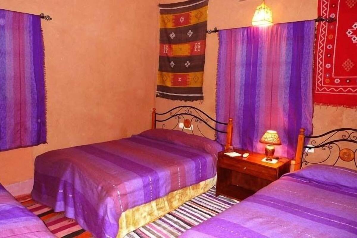 Quarto com duas camas, cada uma com colcha roxa, mesa de cabeceira de madeira com abajur e tapete estampado no chão.