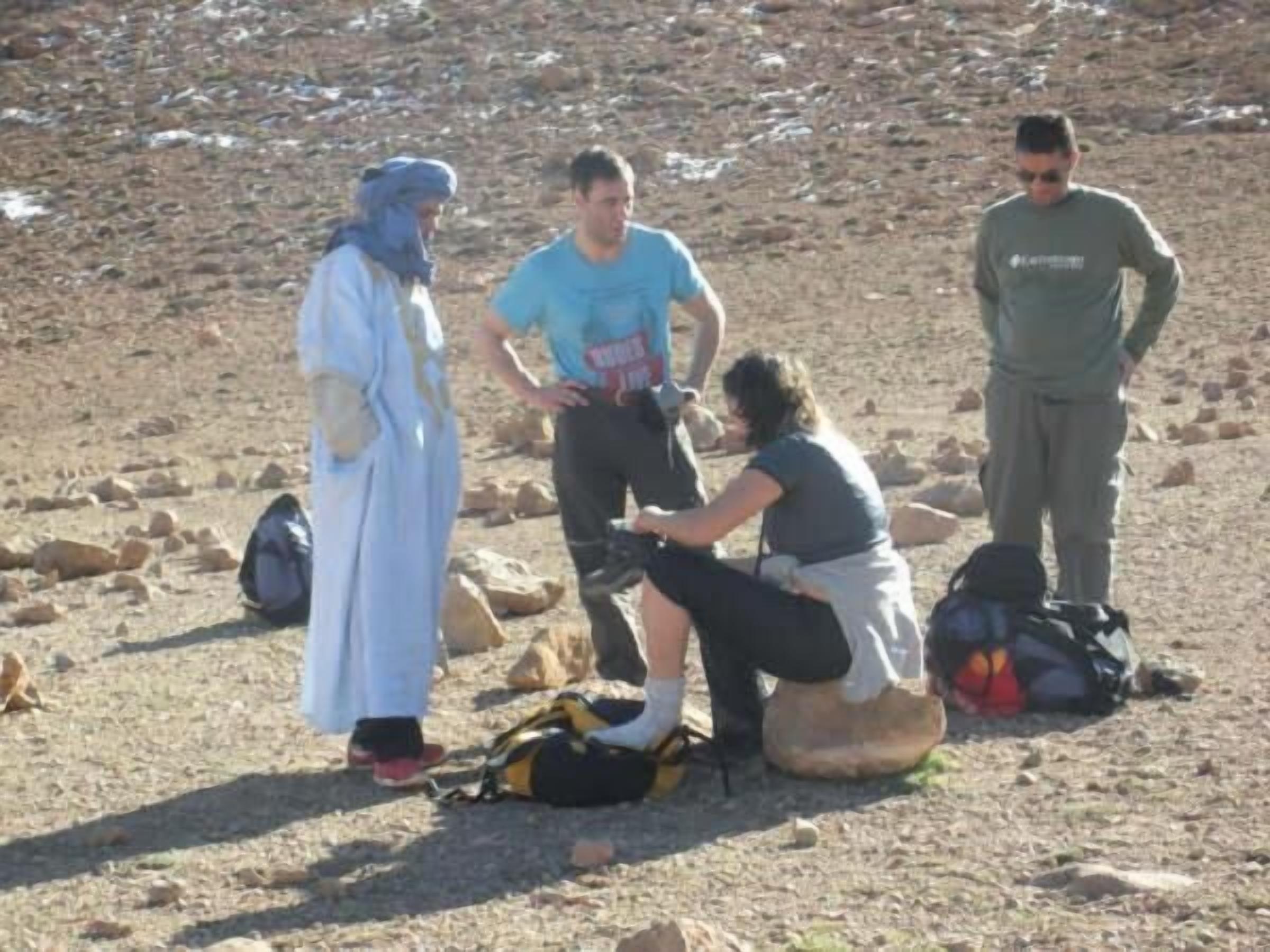 Un grupo de personas al aire libre, algunas sentadas y otras de pie, con mochilas y un bolso en el suelo.