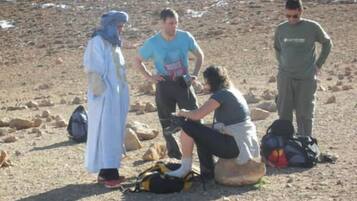 Un grupo de personas al aire libre, algunas sentadas y otras de pie, con mochilas y un bolso en el suelo.