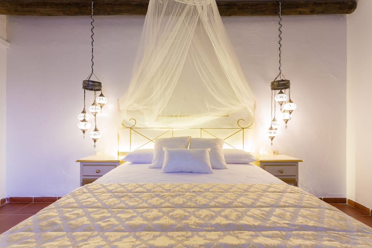 Un dormitorio con una cama grande, ropa de cama blanca y dosel. Hay dos mesitas de noche con lámparas.