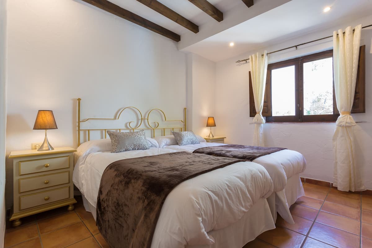 Un dormitorio con dos camas, una mesita de noche con lámpara y una ventana con cortinas.