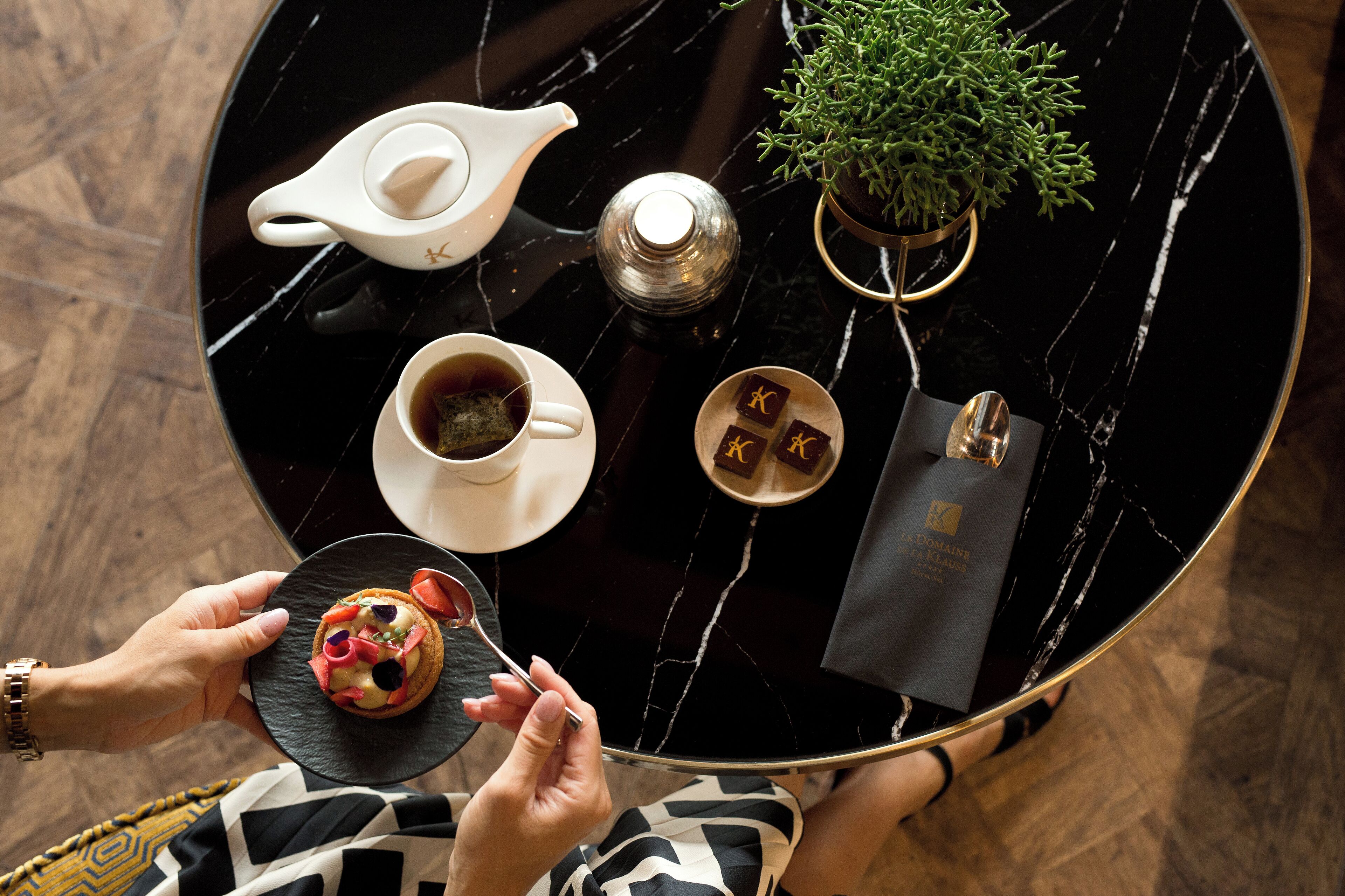 Ein runder, schwarzer Marmor-Tisch mit Teekanne, Tasse Tee, Teller mit Speisen, kleiner Pflanze und Buch.