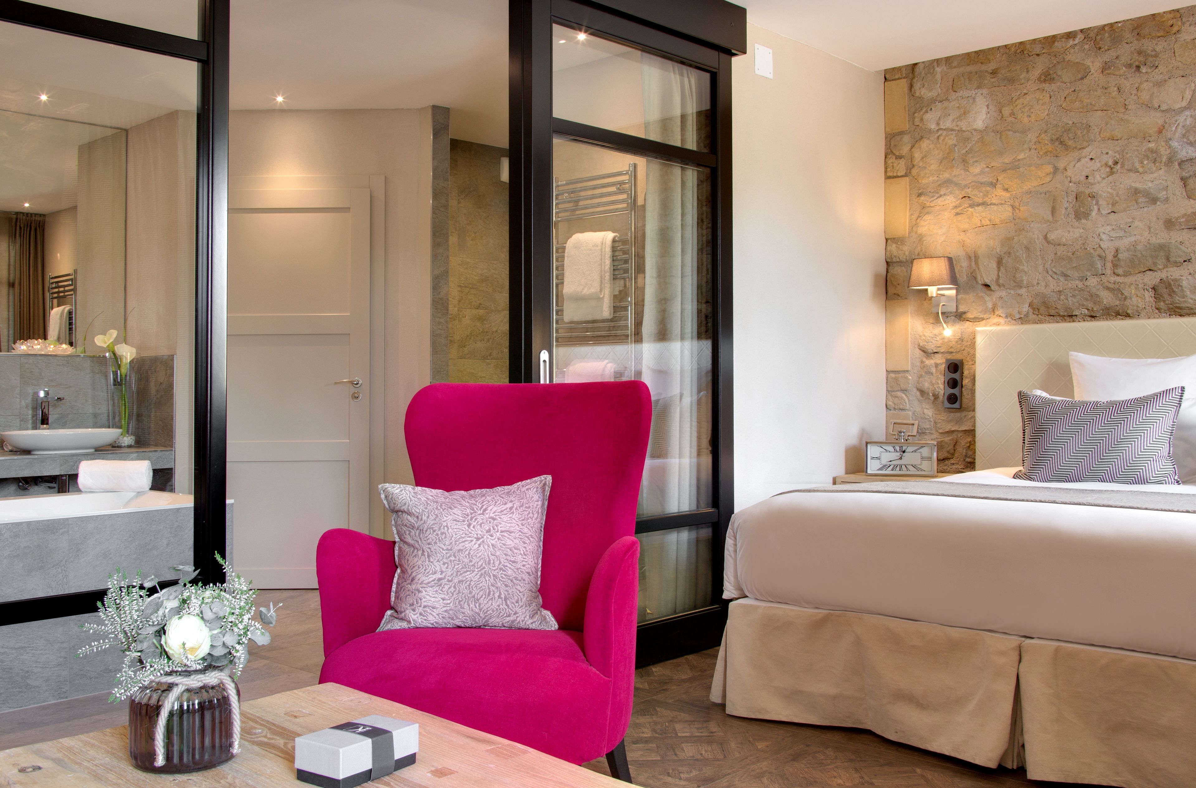 Una habitación de hotel moderna con una cama grande, un sillón rosa brillante, una mesita con un florero y una pared de piedra.