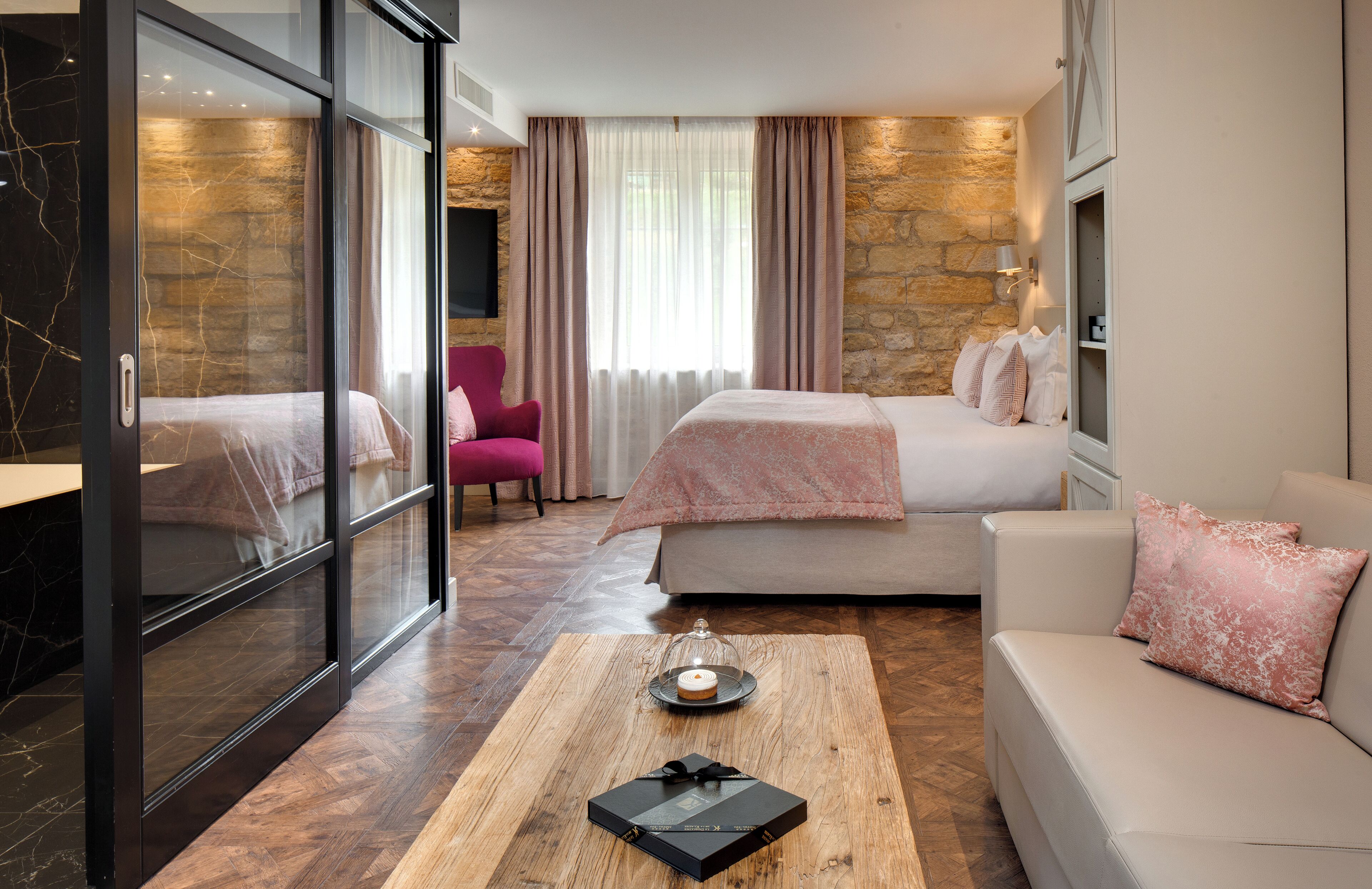 Een moderne hotelkamer met een bed, een bank, een bijzettafeltje met een boek en een glas, en een schuifdeur naar een badkamer.