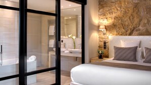 Een moderne slaapkamer met een groot bed, een accentmuur van steen, een badkamer met een glazen deur en een nachtkastje met een lamp.