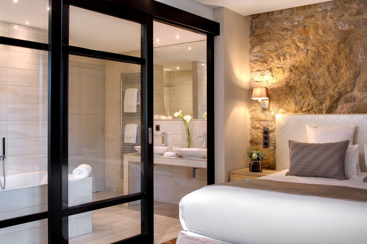 Een moderne slaapkamer met een groot bed, een accentmuur van steen, een badkamer met een glazen deur en een nachtkastje met een lamp.
