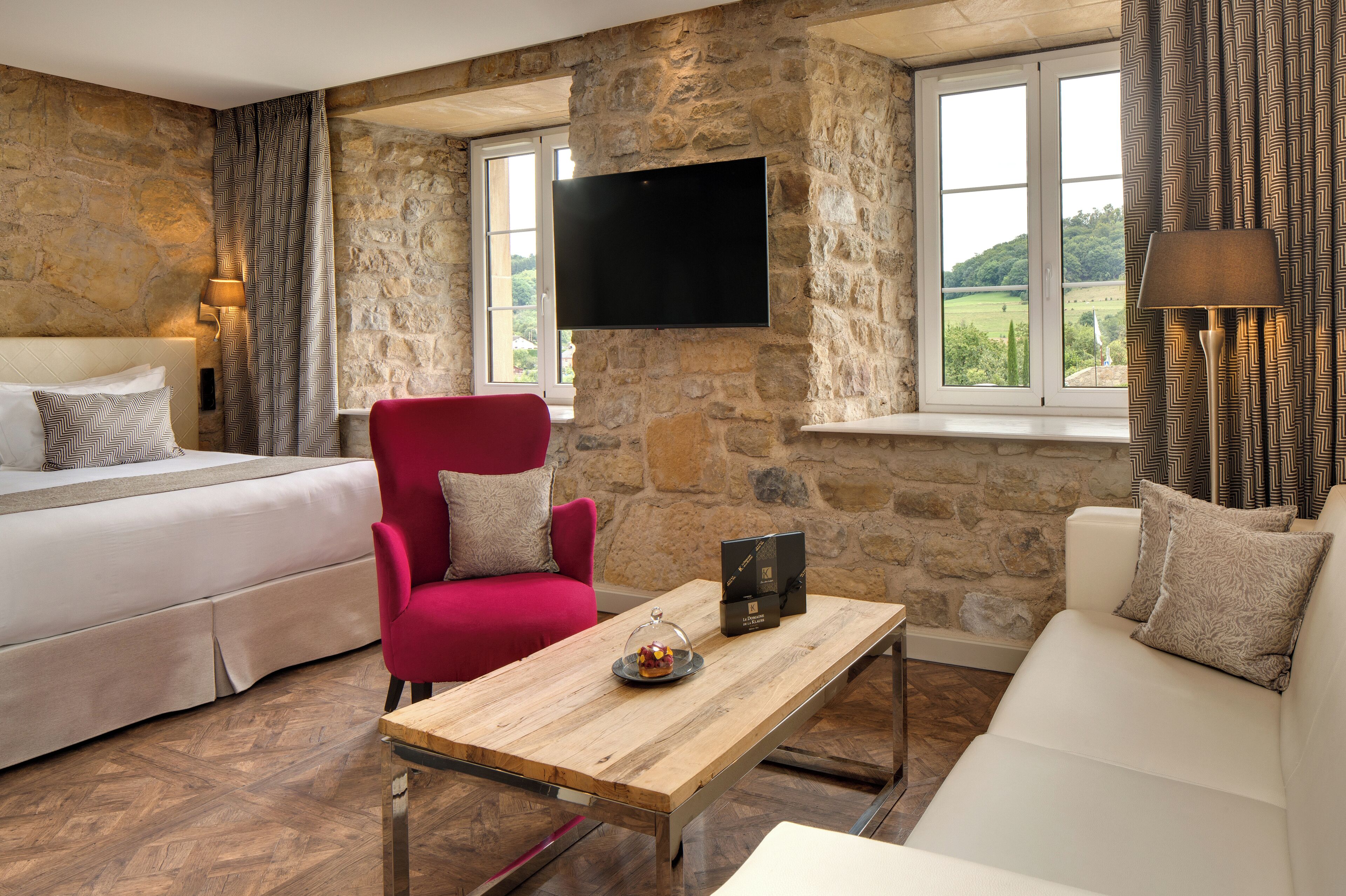 A room with a bed, a red chair, a wooden table, a sofa, a stone wall, and a TV mounted on the wall.
