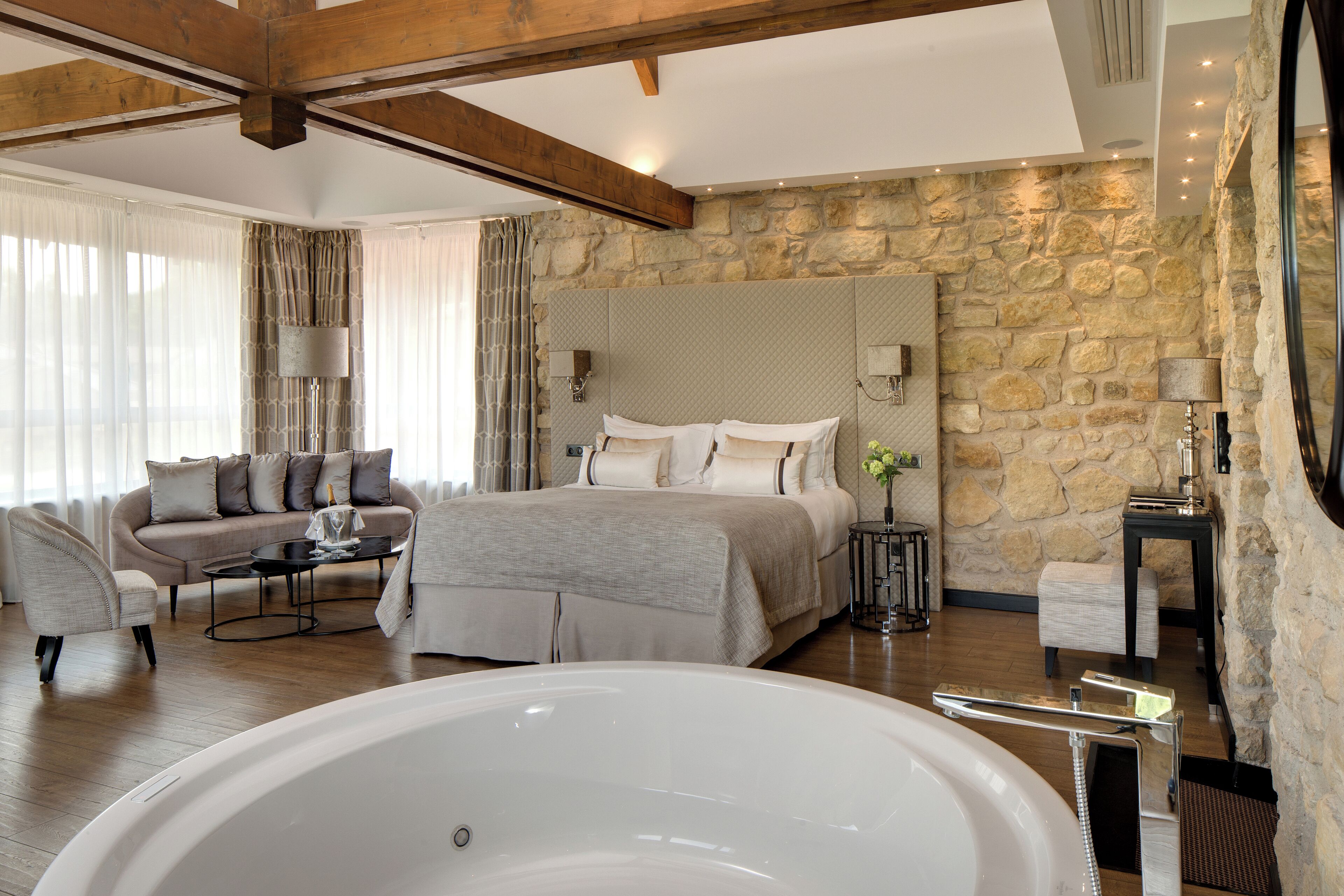 A modern bedroom with a large bed, a sitting area with a sofa and armchairs, a stone accent wall, and a large bathtub.