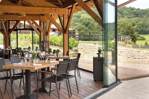 An outdoor dining area with wooden tables and chairs, a stone wall, and a view of a lush green landscape.
