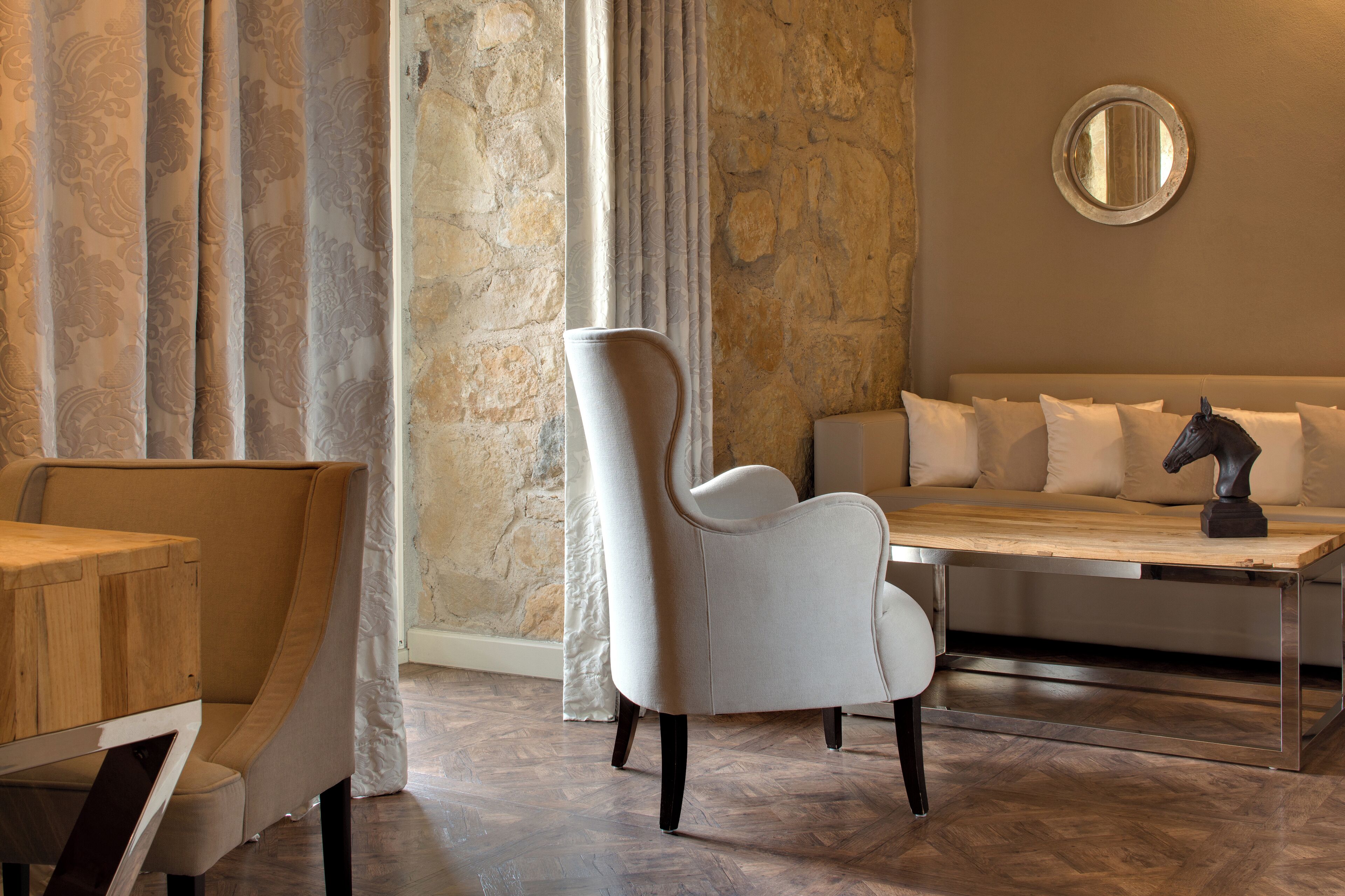 A modern room with a stone wall, a round mirror, a sofa with cushions, a white armchair, and a wooden table.