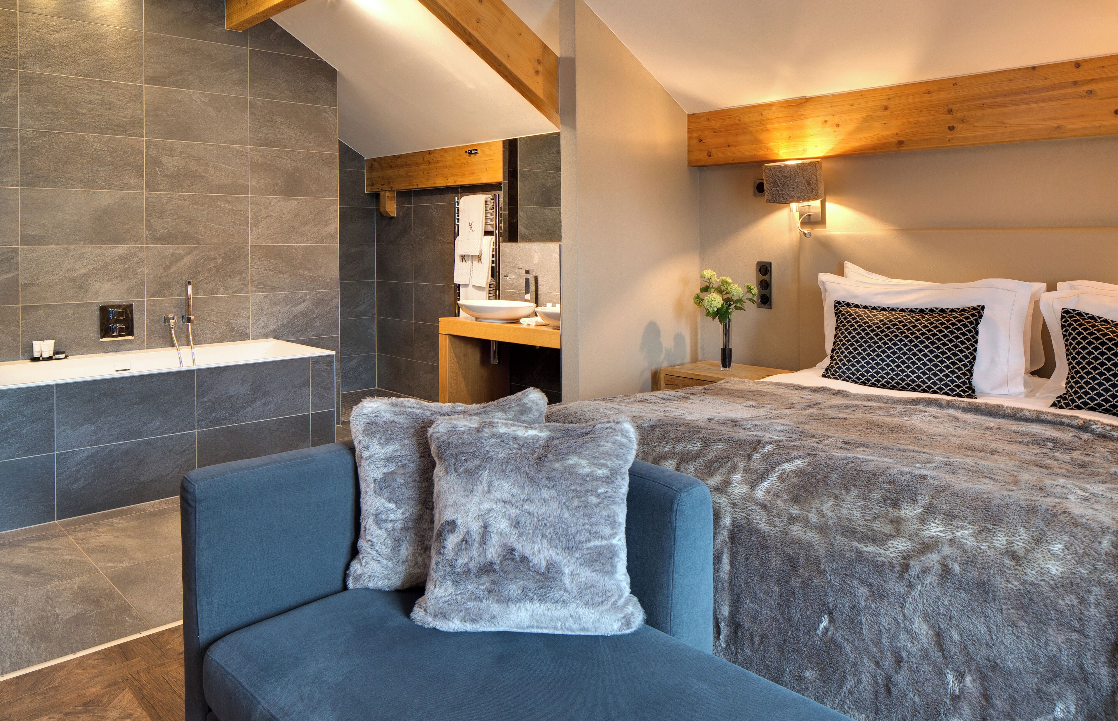 A modern bedroom with a large bed, a sofa, a bathroom with a bathtub, and a wooden ceiling.