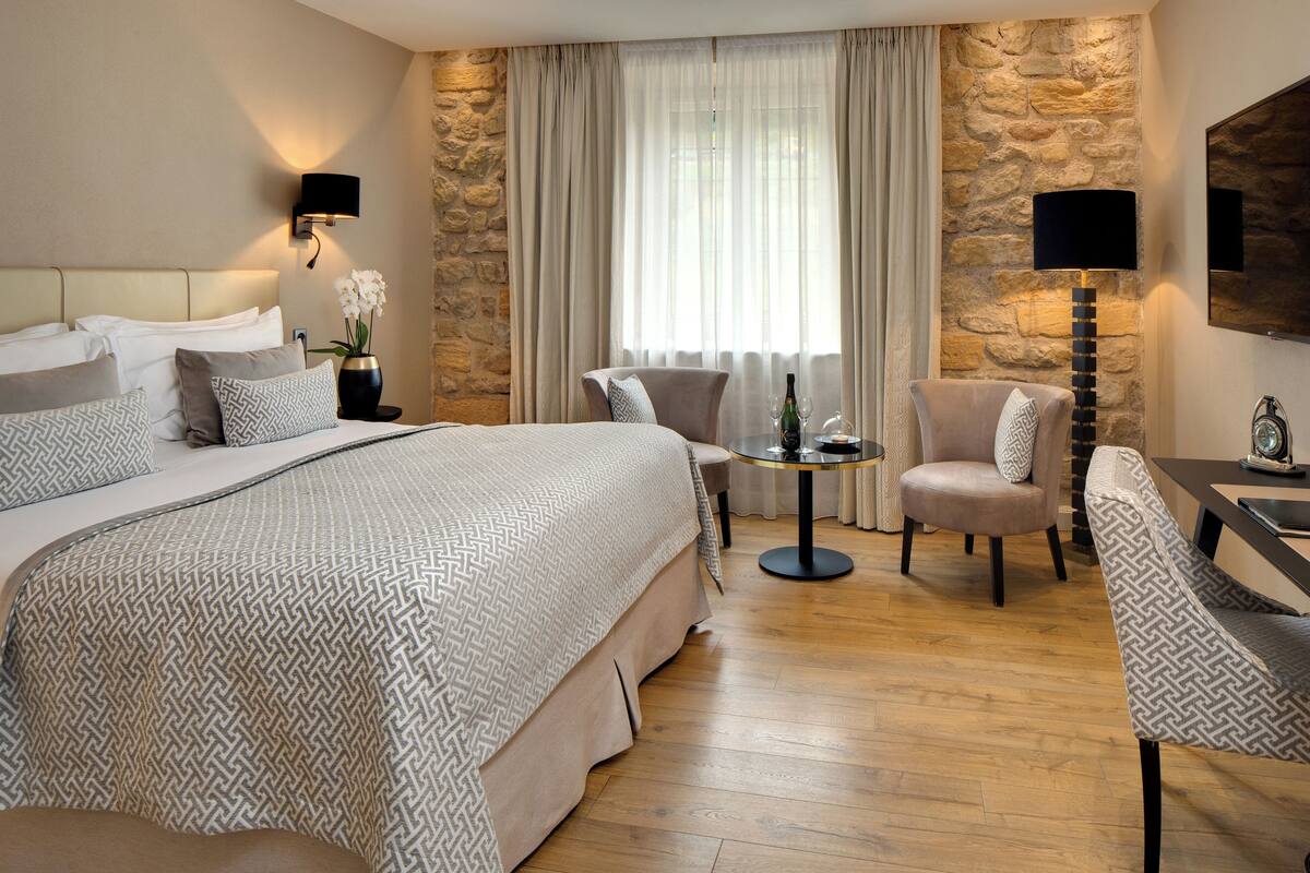 Een hotelkamer met een groot bed, een zithoek met een tafel en stoelen, een accentmuur van steen, en een groot raam met gordijnen.