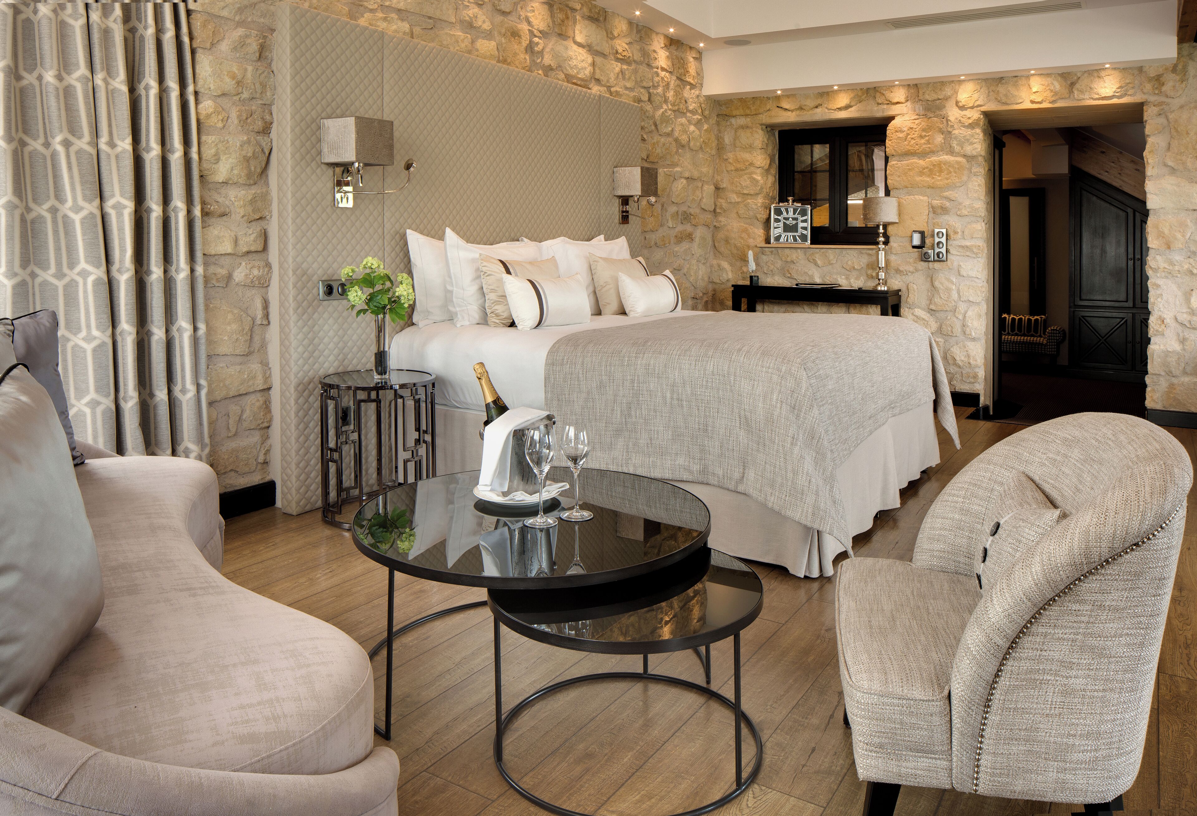 Una habitación de hotel moderna con una cama grande, una mesa redonda de vidrio, dos sillones y una pared de acento de piedra.