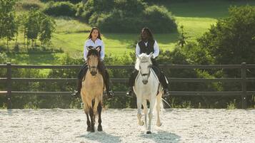 Two people riding horses in an outdoor arena with a wooden fence and green fields in the background.