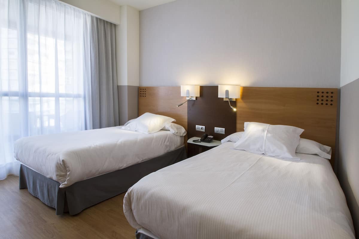 Camera d'albergo con due letti, una testiera in legno, lampade ai comodini e una finestra con tende.