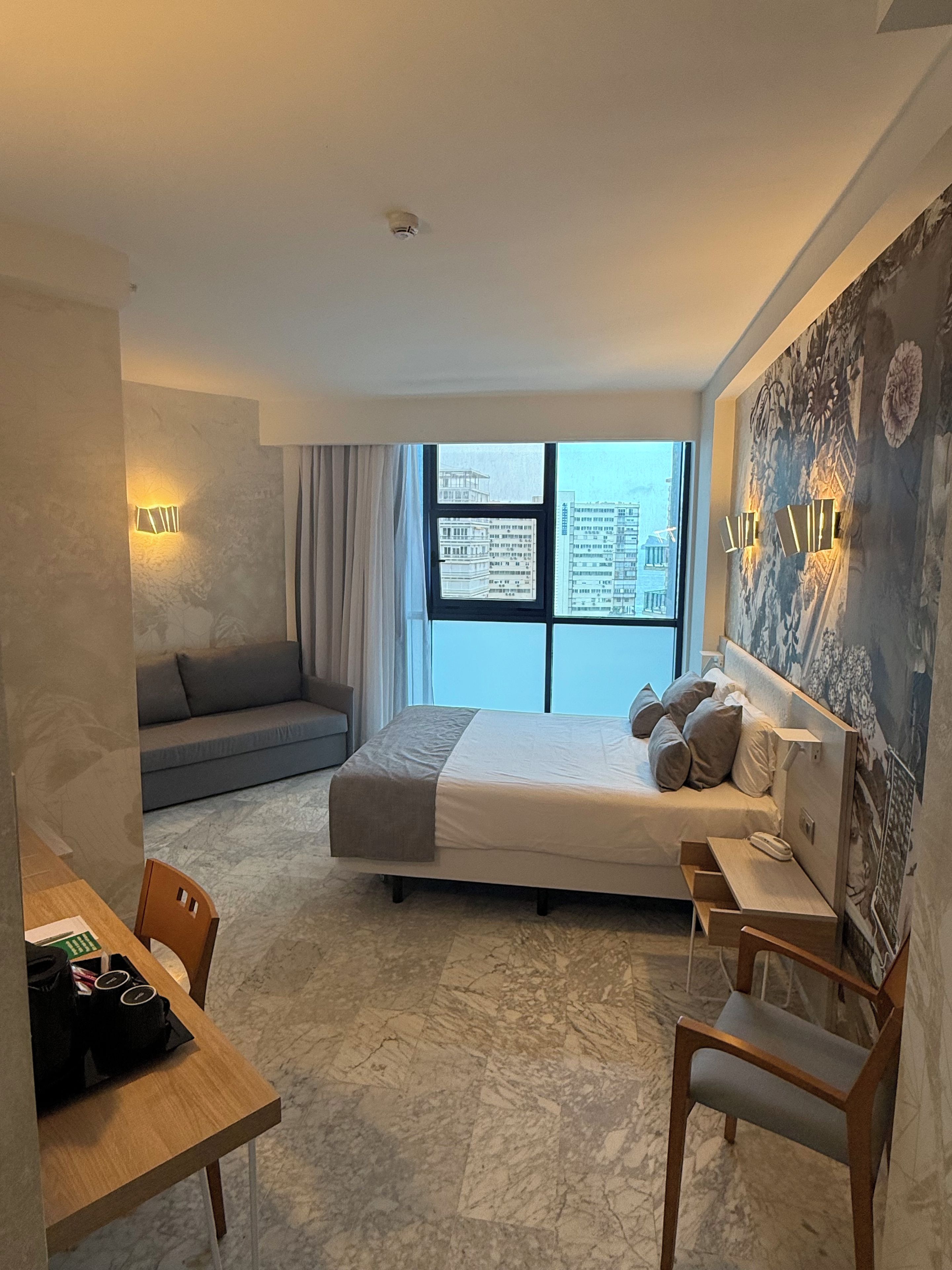 A modern hotel room with a large bed, a desk, a sofa, and a view of buildings through the window.