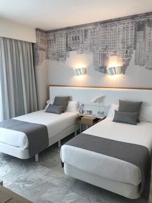 Camera d'albergo con due letti, un murale raffigurante un panorama urbano, un comodino e una lampada.