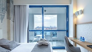 A hotel room with a bed, a desk with a chair, a table with two bottles, and a view of a cityscape through a large window. - Madeira Centro (Benidorm)