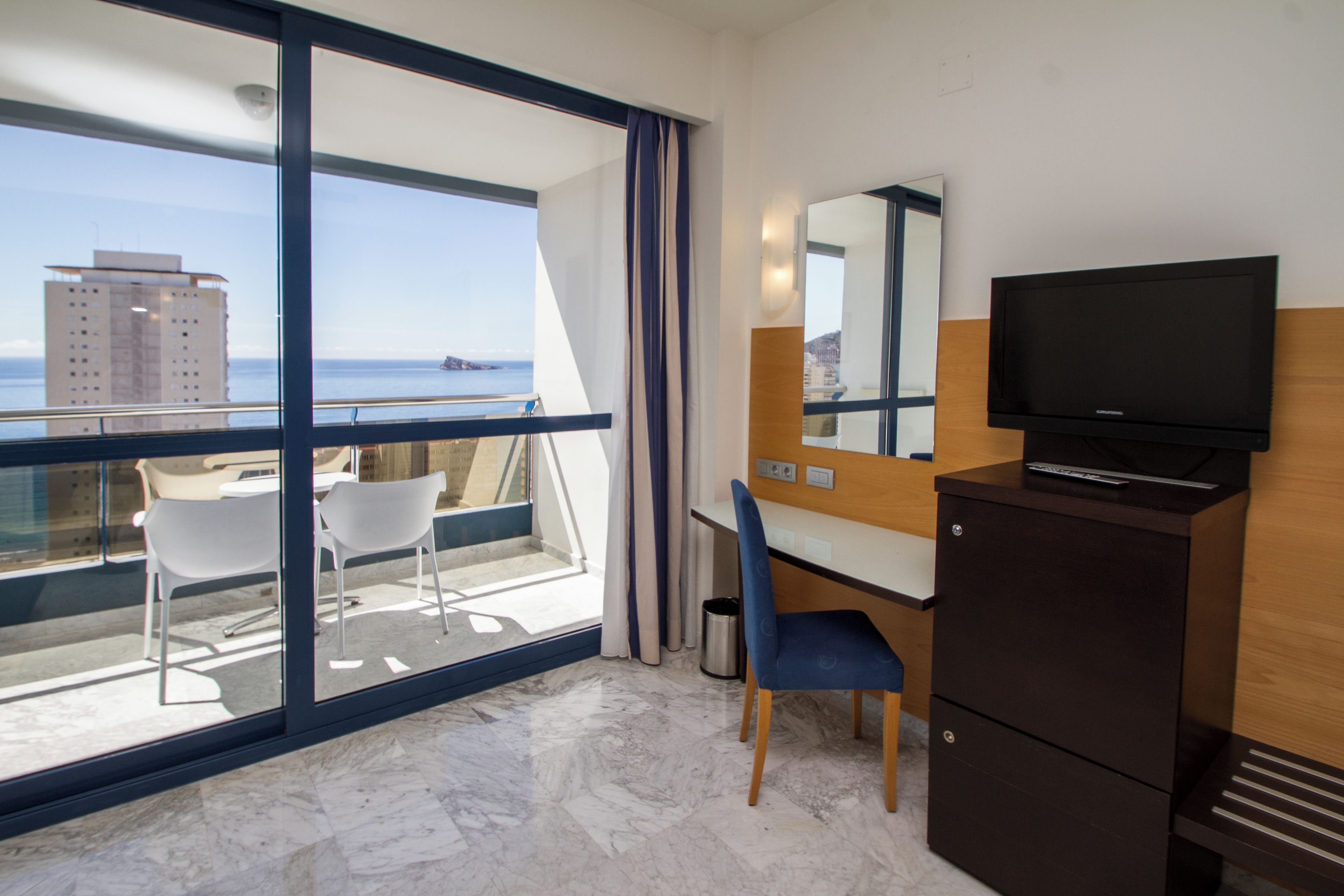 Habitación de hotel con un ventanal grande, escritorio, silla, televisor y minibar.