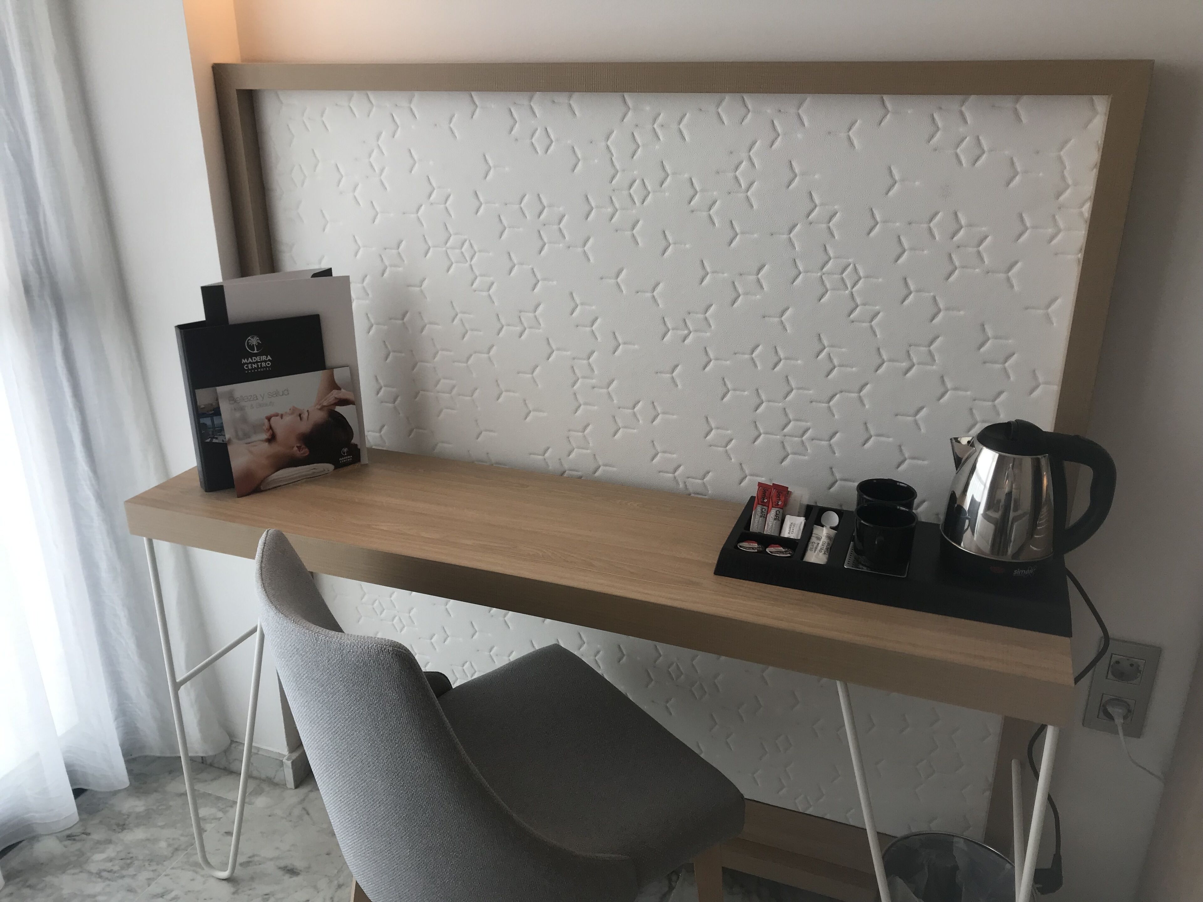 Een hotel bureau met een houten blad, een stoel, een witte structuurwand en een klein zwart dienblad met een waterkoker en andere spullen.