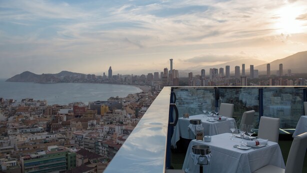 A rooftop dining area with tables set for two, overlooking a coastal cityscape with a clear view of the sea and a distant mountain range. - Madeira Centro (Benidorm)