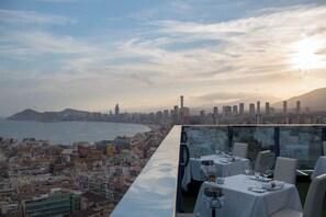 Zona pranzo sul tetto con tavoli apparecchiati per due, con vista sulla città costiera e una limpida visuale del mare e di una catena montuosa in lontananza.