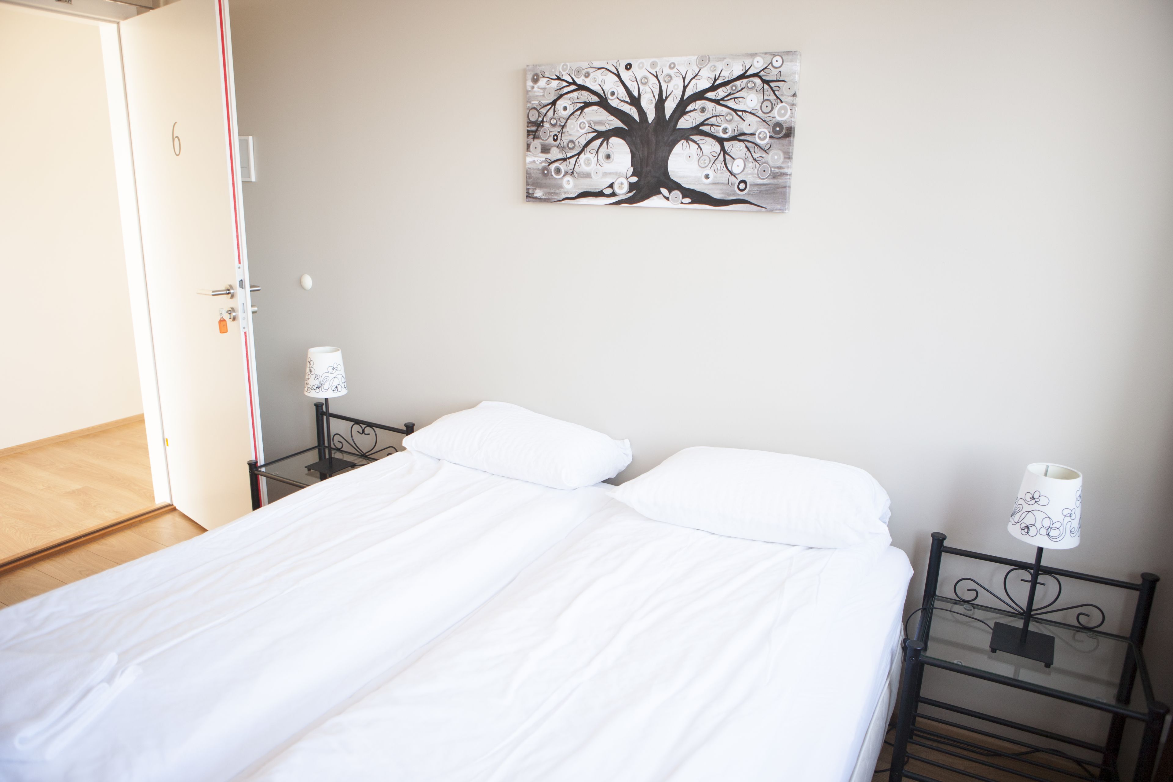 A bedroom with a bed, two bedside tables with lamps, a wall-mounted painting of a tree, and a door.