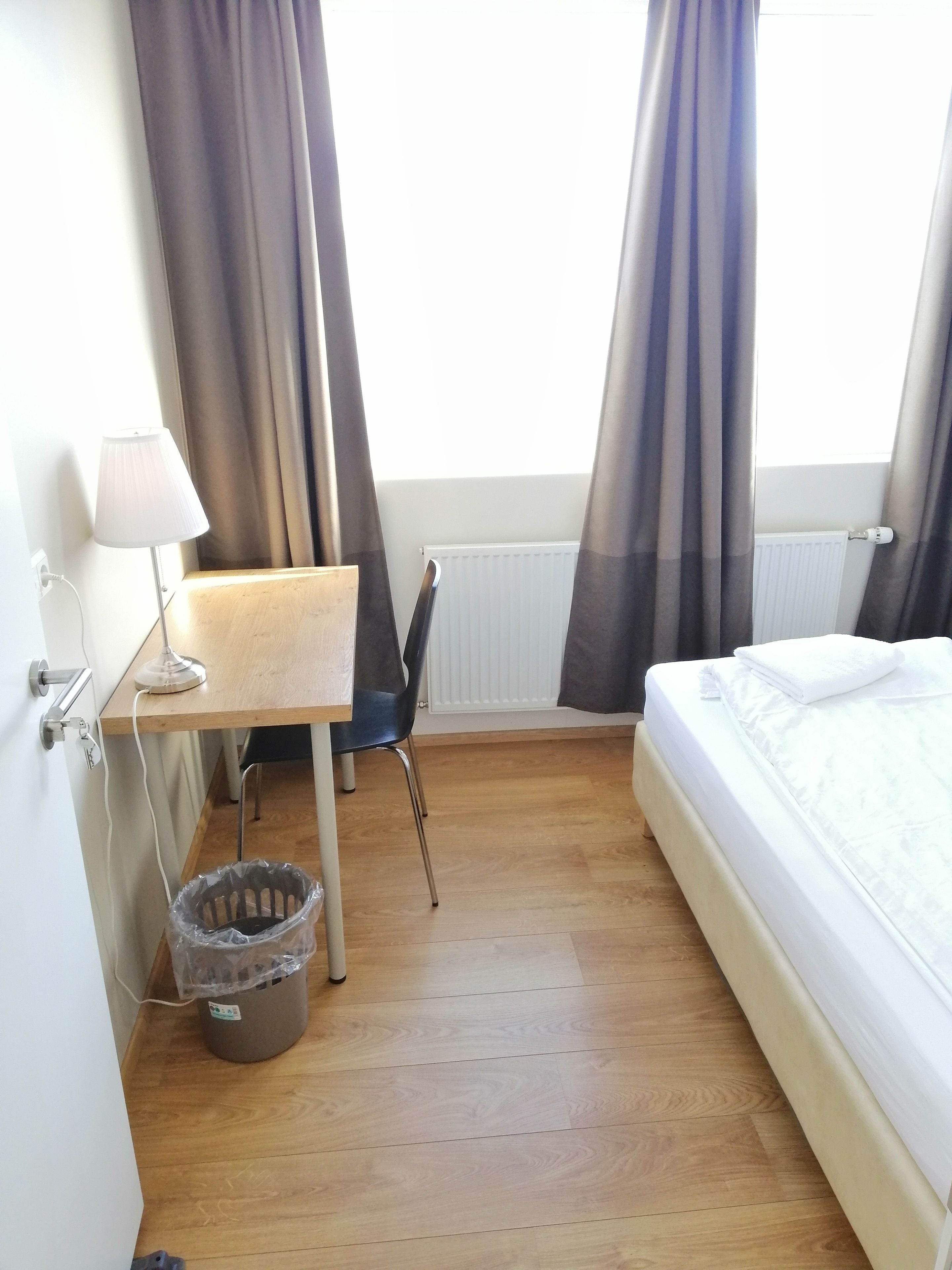 Una habitación de hotel pequeña con suelo de madera, un escritorio con lámpara, una silla, una cama con ropa blanca y un radiador.