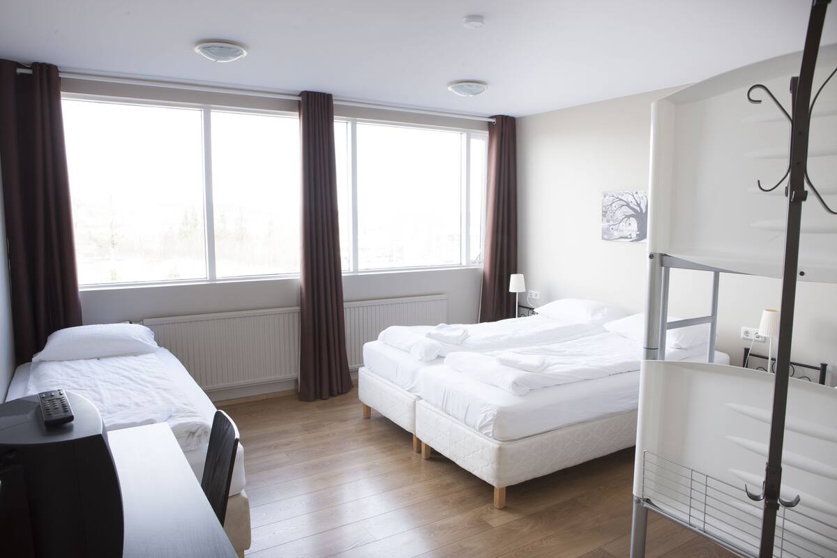 Ett hotellrum med två sängar, ett skrivbord, en stol, ett fönster med gardiner, ett element och en hylla som är monterad på väggen.