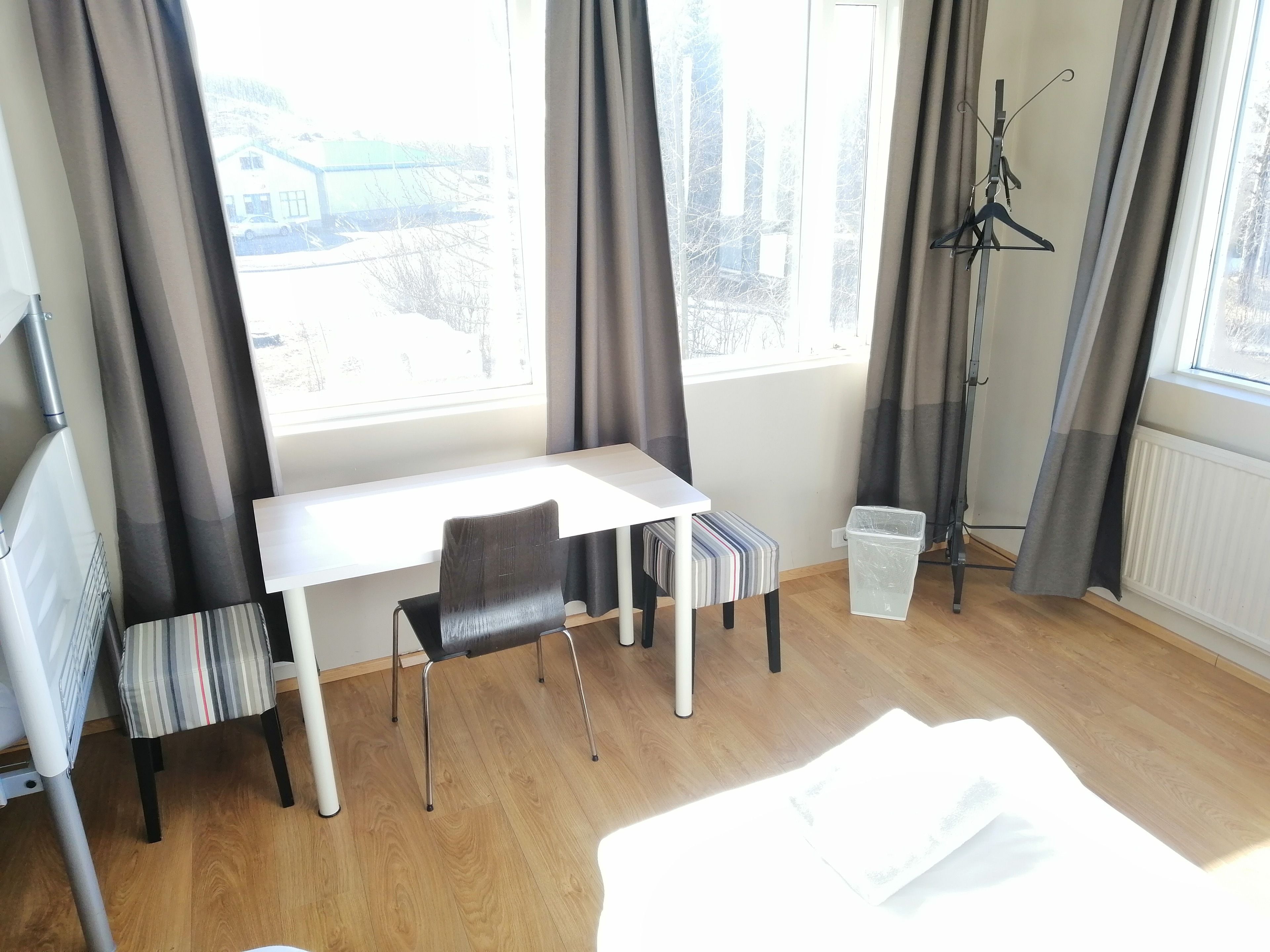 A hotel room with a white desk, a black chair, a striped ottoman, a radiator, and a window with curtains.