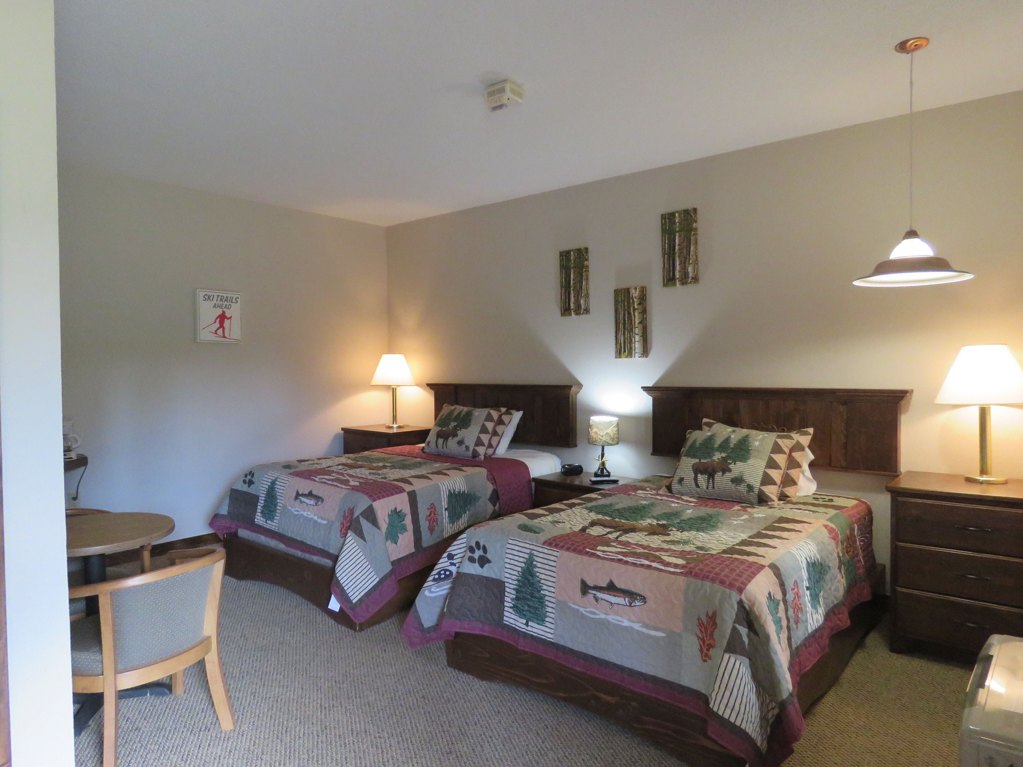 A hotel room with two beds, a small table, and a chair. There are wall-mounted lamps and a ceiling light.