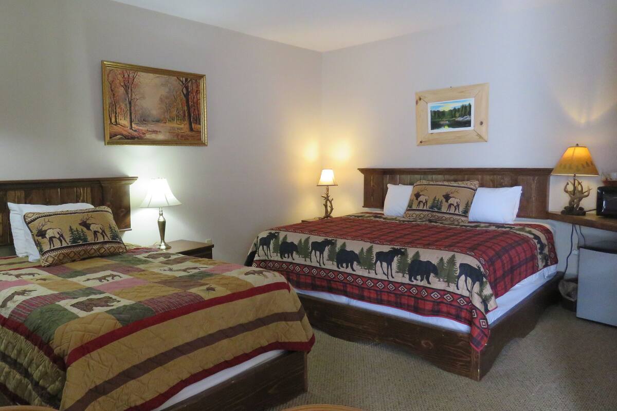 A hotel room with two beds, each with a unique quilt, a wooden headboard, bedside lamps, and a painting on the wall.