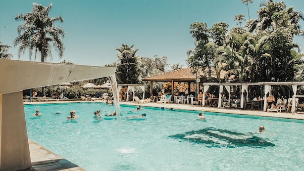 Una piscina con gente nadando y tomando el sol, rodeada de palmeras y zonas con sombra.
