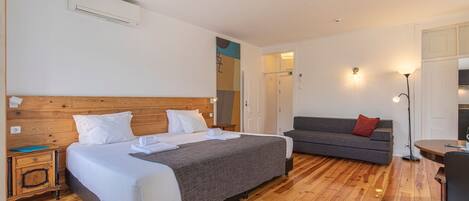 Ein Hotelzimmer mit Holzboden, einem Doppelbett mit weißer Bettwäsche, einem grauen Sofa, einem kleinen Tisch und einer Wand-Klimaanlage.