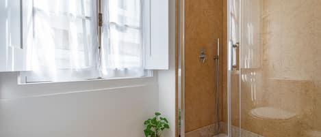 Un baño con mampara de vidrio, un lavamanos blanco, una planta en maceta y una toalla colgada en la pared.