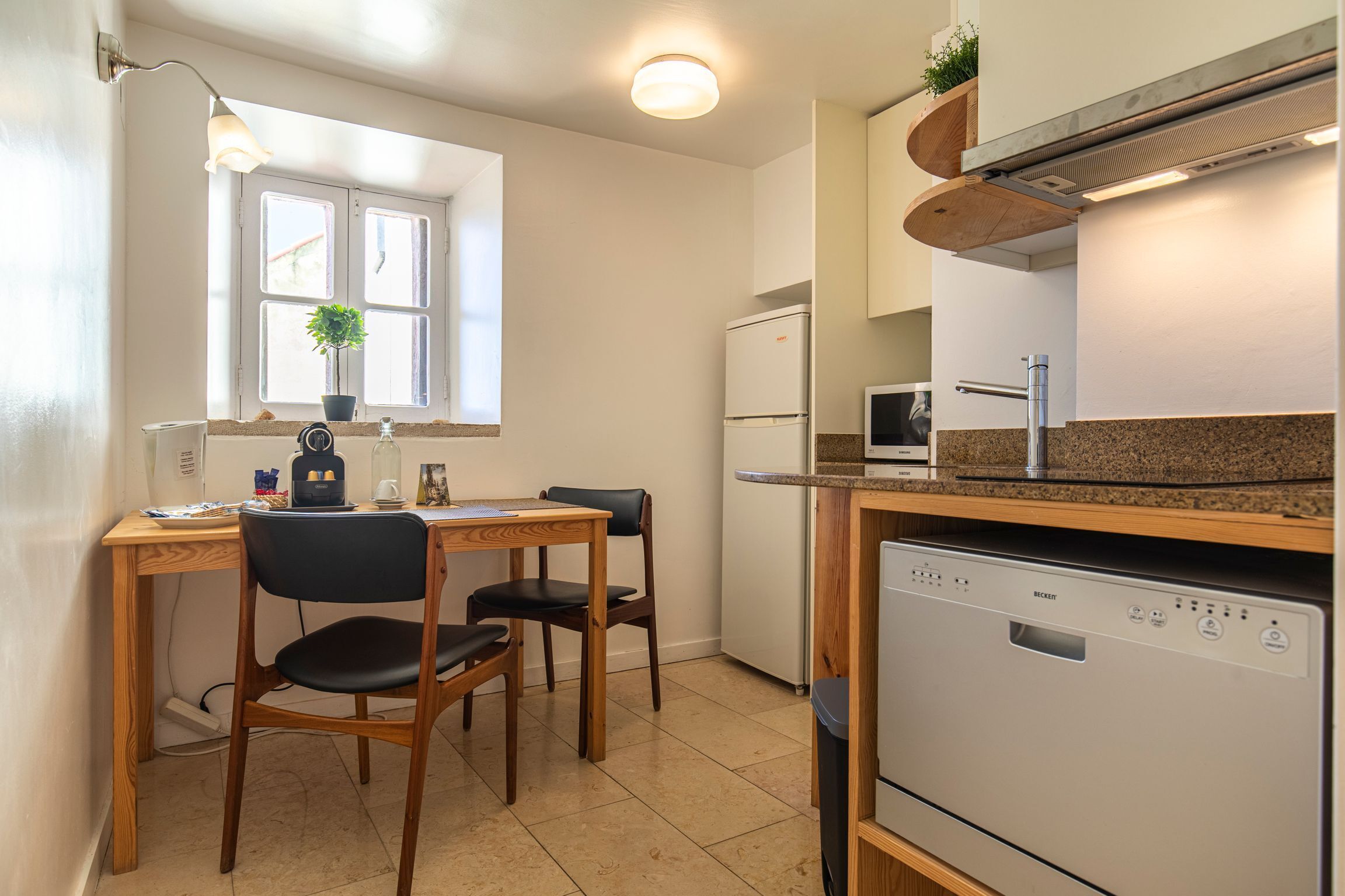 Une cuisine compacte comprenant une table à manger, des chaises, un réfrigérateur, un lave-vaisselle et une fenêtre agrémentée d’une plante.