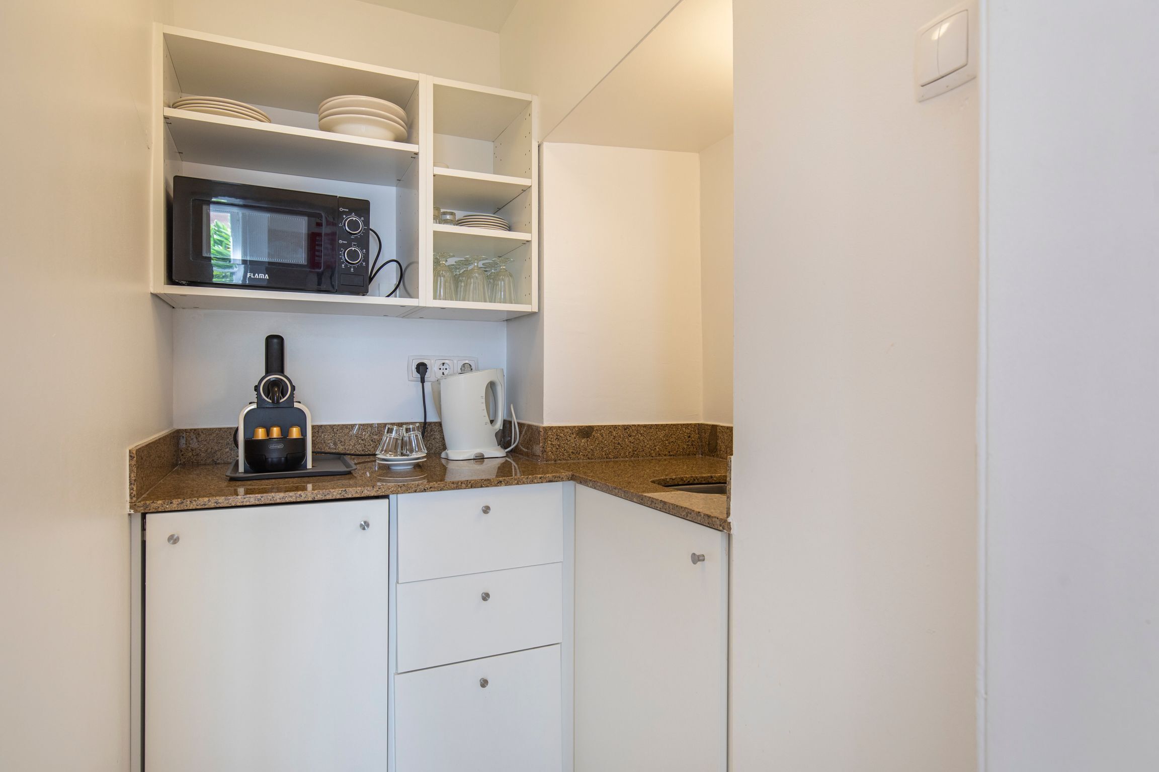 Cocina compacta con armarios blancos, microondas, cafetera y lavabo.