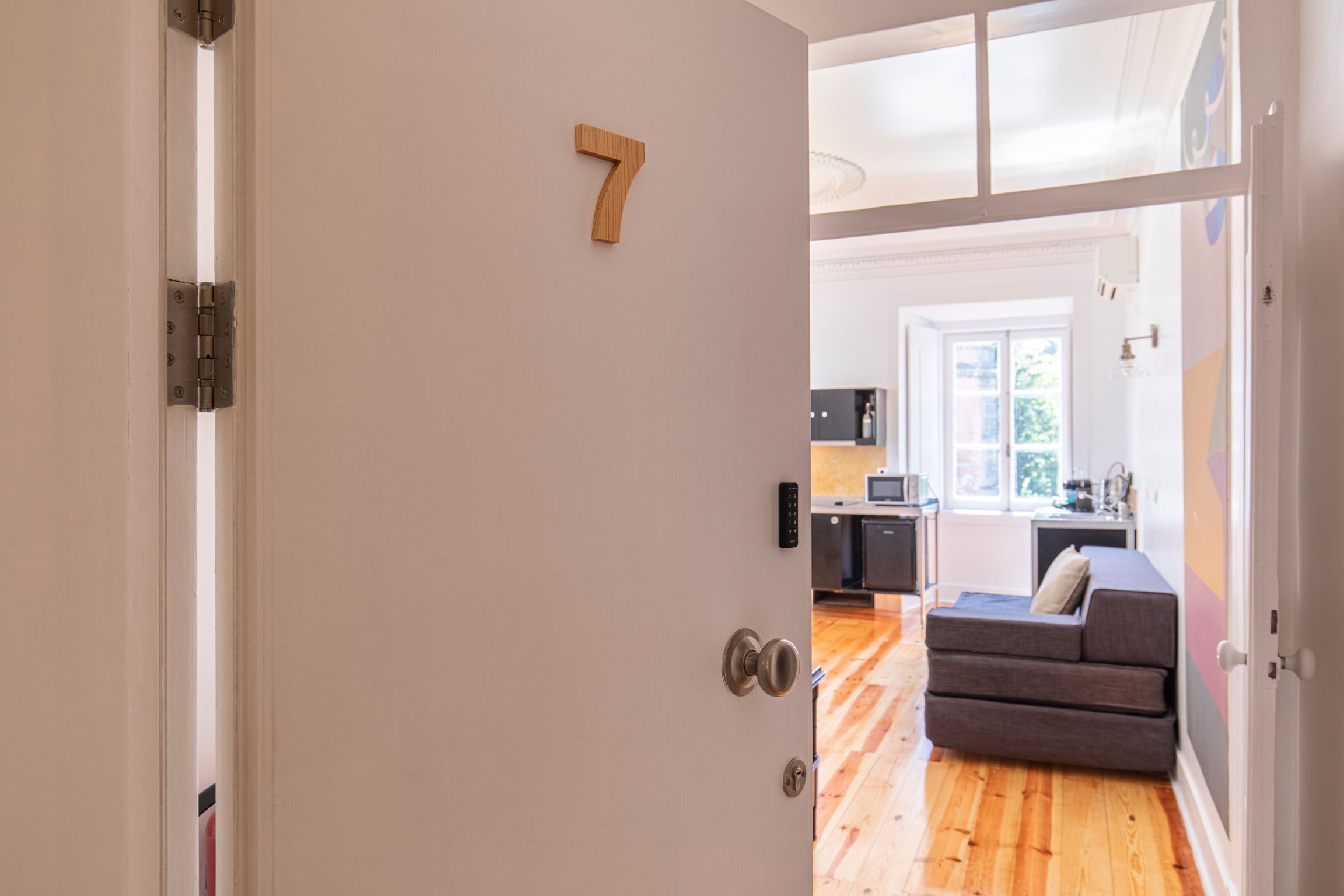 Un interior moderno con una puerta de madera marcada con el número 7, que da paso a una sala de estar con un sofá y una cocina al fondo.