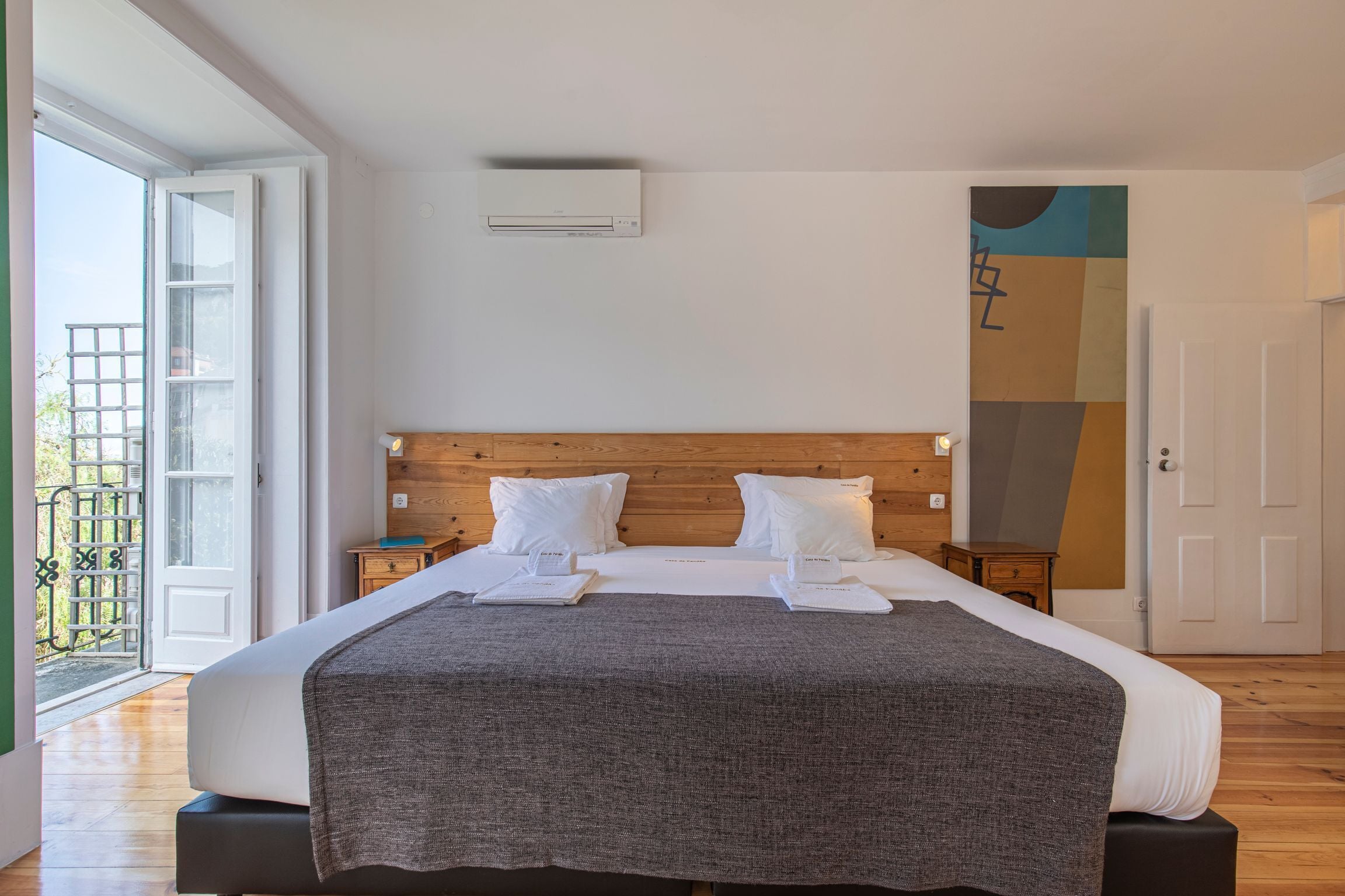 Un lit bien fait, avec du linge de lit blanc et un plaid gris, une tête de lit en bois et un grand miroir au mur.