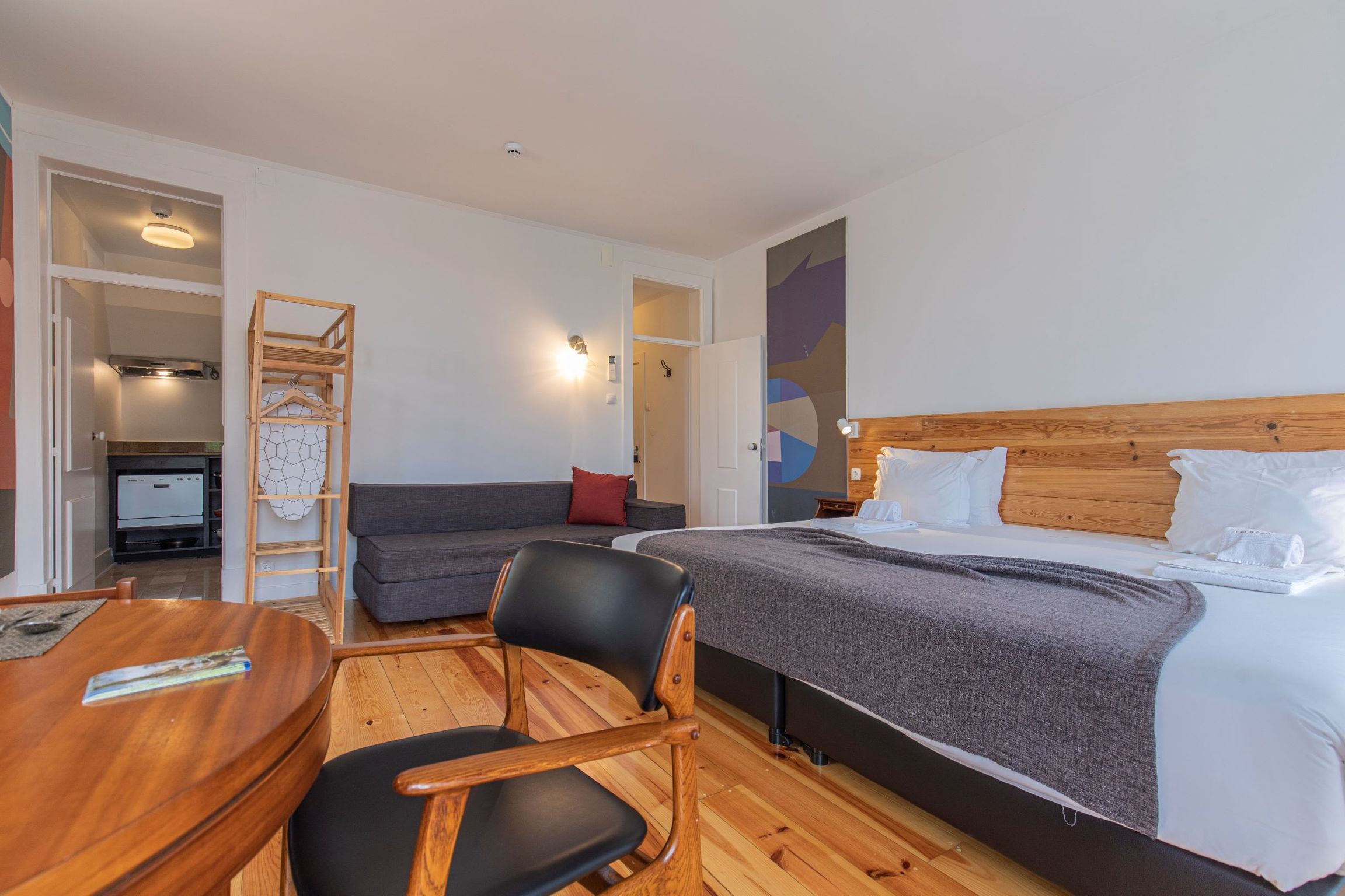 A hotel room with a bed, a sofa, a desk, and a chair. There is a wooden ladder and a shelf with clothes.