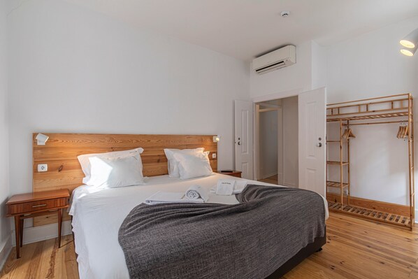 Una cama bien hecha con ropa blanca, cabecera de madera y manta gris. Se observa un mesita de noche de madera con un cajón y una pequeña mesa al lado de la cama. En el fondo, un armario de madera con espacio para colgar.