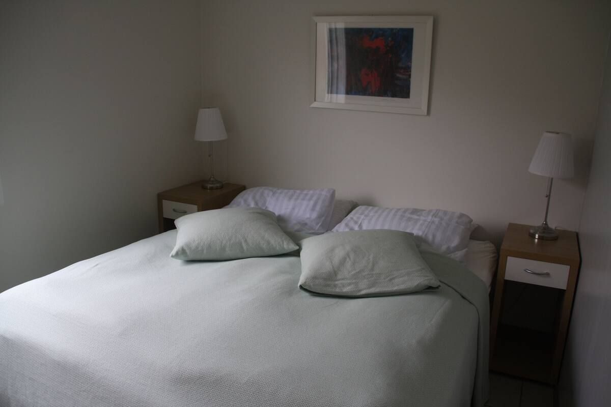 Una cama bien hecha con sábanas blancas y dos almohadas, una mesita de noche con lámpara y un cuadro en la pared.