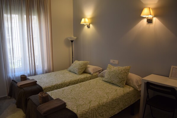 Quarto de hotel com duas camas, uma escrivaninha, uma cadeira, um abajur e uma janela com cortinas.