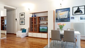 A modern living room with a wooden floor, a white sofa, a dining table with chairs, a TV, and a bookshelf. - Padovaresidence Piazza delle Erbe (Padova)