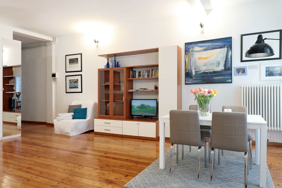 A modern living room with a wooden floor, a white sofa, a dining table with chairs, a TV on a wooden cabinet, and artwork on the walls.