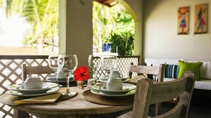 Una mesa de comedor dispuesta con platos, vasos y un centro de mesa con una flor roja, rodeada por una silla de madera y un sofá blanco con cojines verdes.