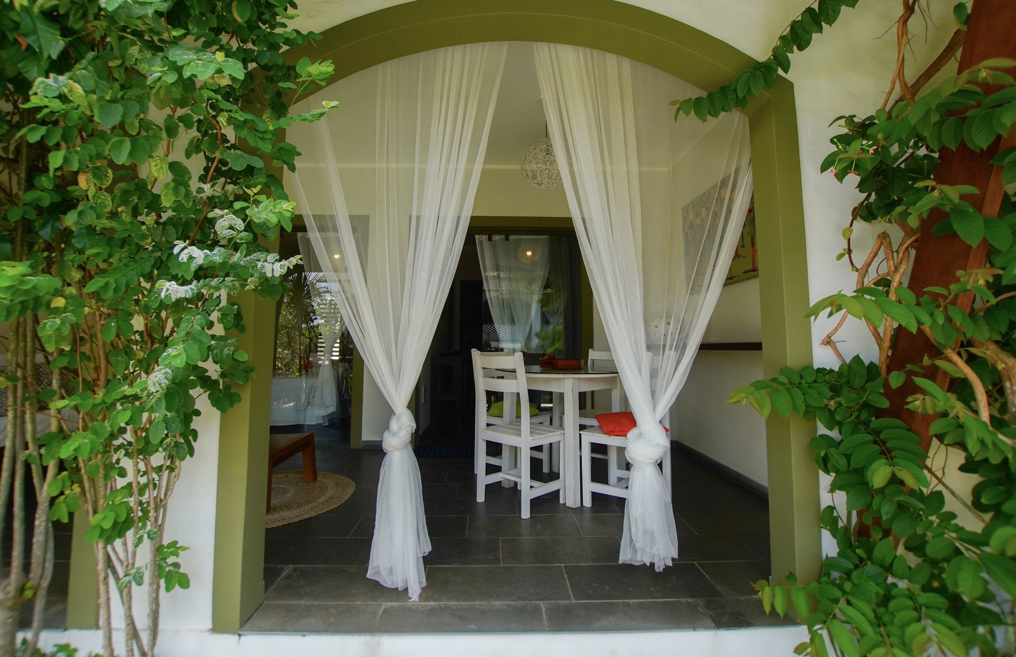An arched doorway with white curtains, a white table with chairs, and green foliage.