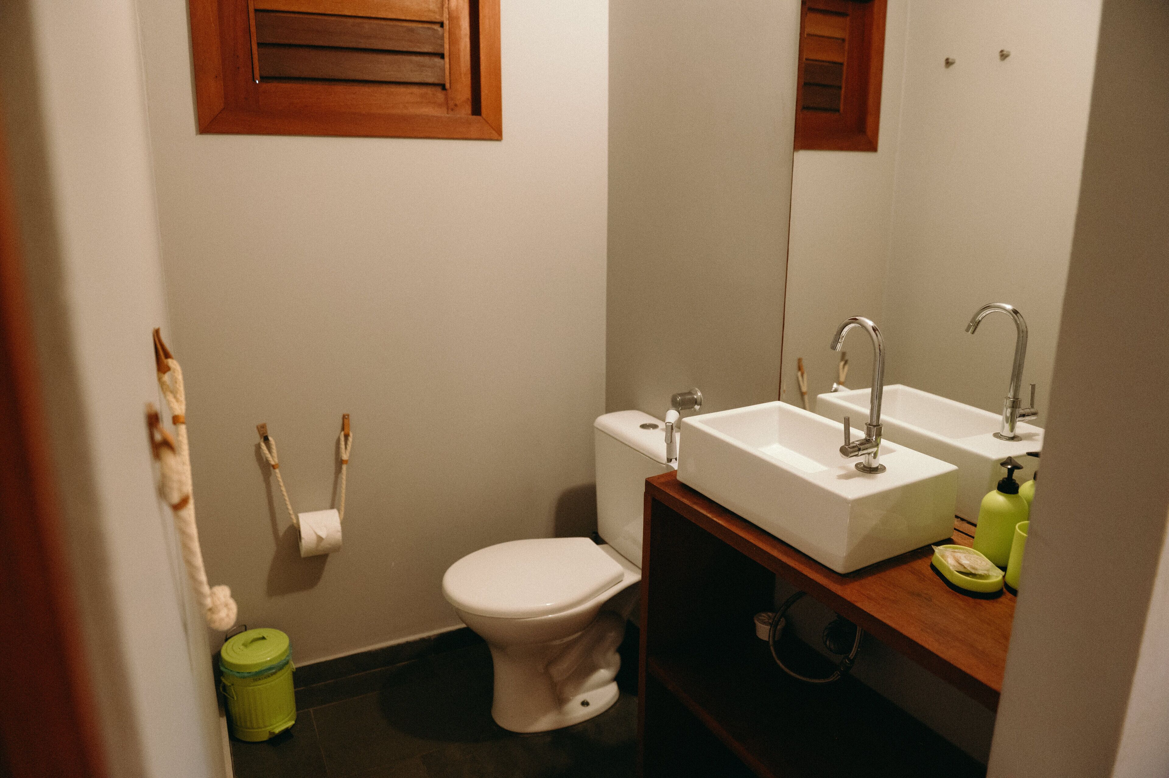 A small bathroom with a toilet, two sinks, a mirror, and a green trash can.