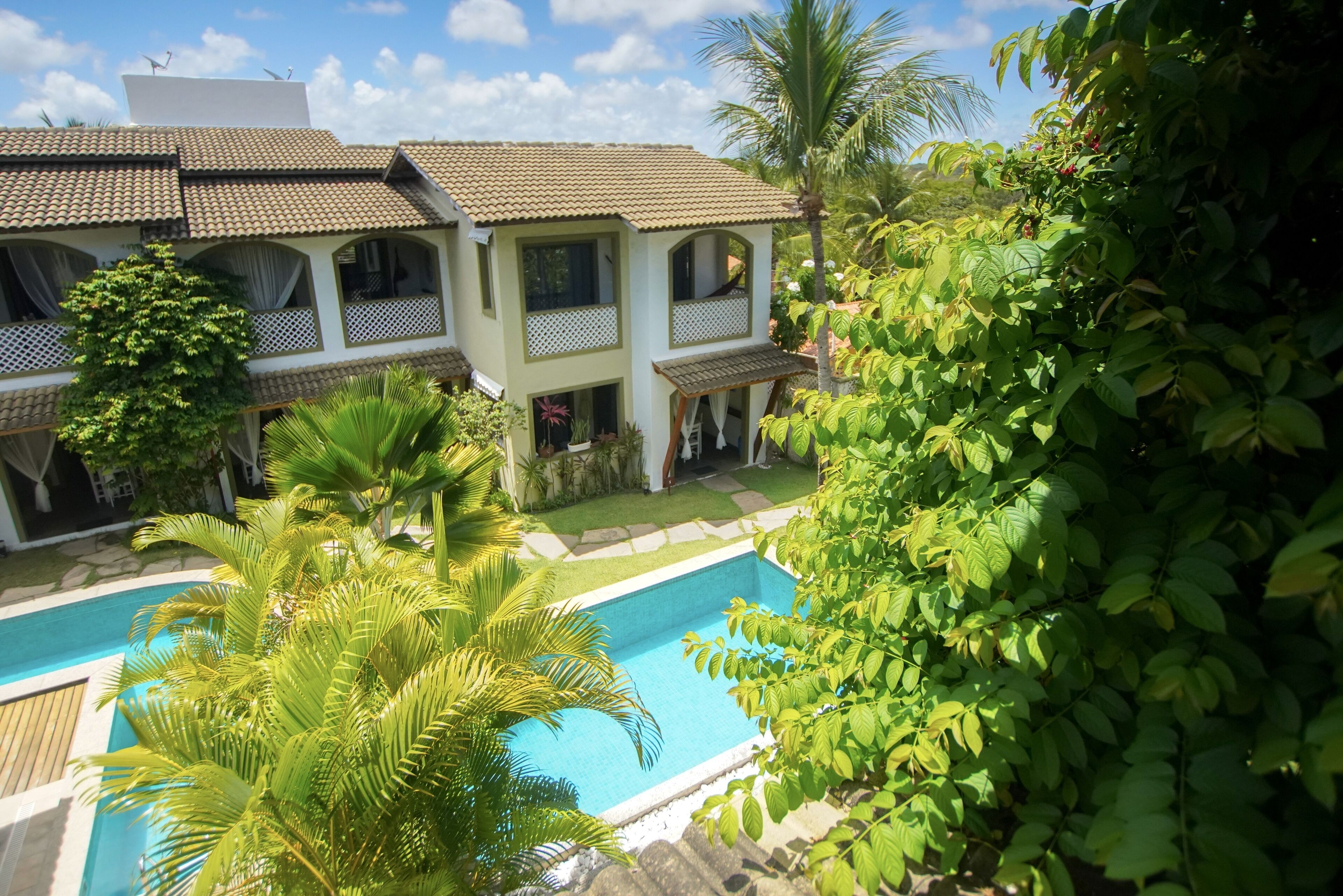 Uma casa de dois andares com piscina, rodeada por vegetação exuberante e palmeiras.