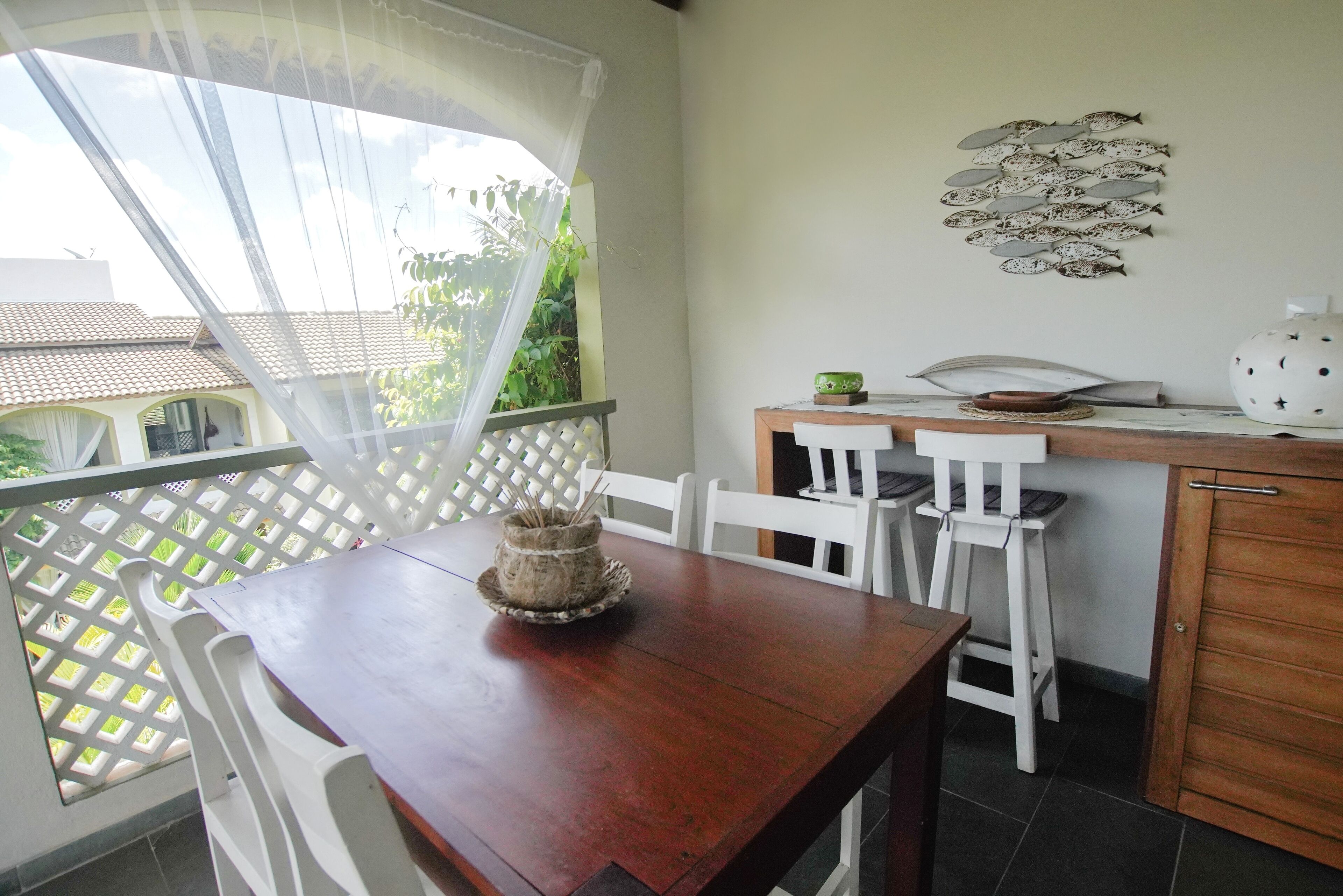 Un comedor con mesa de madera y sillas blancas, una pequeña cocina con lavamanos y alacenas, y un adorno de pared con peces.